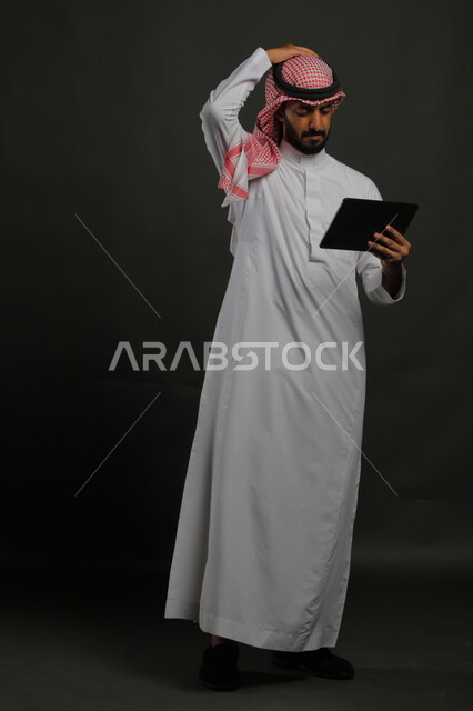 تعابير الانزعاج بتلقي الأخبار السيئة، استخدام التكنولوجيا والتقنية الحديثة، بورتريه لرجل عربي خليجي سعودي يرتدي الزي التقليدي يحمل بيده جهاز التابلت ويضع يده على رأسه بإيماءات خسارة المنتخب المفضل في المباراة، صورة الجسم بالكامل, خلفية سوداء
