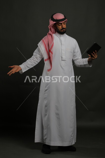 عقد اجتماع عمل عن بعد باستخدام جهاز التابلت، الدردشة مع الأهل والأصدقاء عبر الأونلاين، استخدام التقنيات الحديثة والمطورة، بورتريه لرجل عربي خليجي سعودي يرتدي الزي التقليدي يفتح يده بإيماءات عدم معرفة أمر ما، صورة الجسم بالكامل, خلفية سوداء