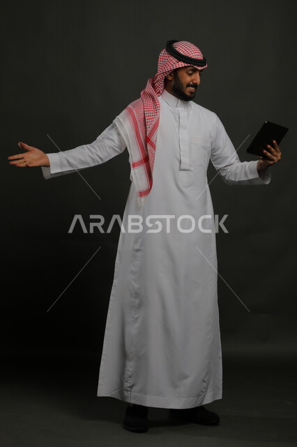 استخدام التقنيات والأجهزة اللوحية الحديثة، الدردشة مع الأهل والأصدقاء عبر الأونلاين، بورتريه لرجل عربي خليجي سعودي مبتسم يرتدي الزي التقليدي يقوم بإجراء مكالمة فيديو باستخدام جهاز التابلت بإيماءات السرور، صورة الجسم بالكامل, خلفية سوداء