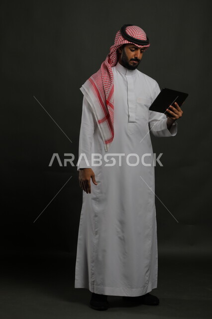 إنجاز مهام العمل عن بعد، متابعة آخر الأخبار على مواقع الانترنت، دمج التكنولوجيا والتقنية الحديثة بالحياة العملية، بورتريه لرجل عربي خليجي سعودي يرتدي الزي التقليدي يحمل بيده جهاز التابلت، صورة الجسم بالطول الكامل, خلفية سوداء