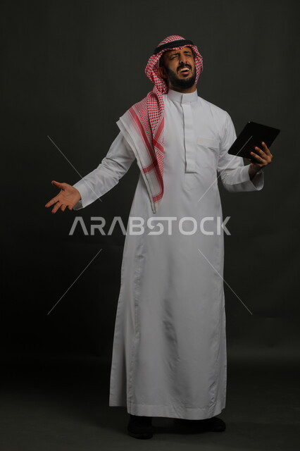 تشجيع المنتخب الوطني السعودي، إيماءات الانزعاج والغضب لخسارة فريق كرة القدم المفضل في المباراة, بورتريه لرجل عربي خليجي سعودي يرتدي الثوب التقليدي والشماغ يحمل بيده جهاز التابلت، استخدام التكنولوجيا والتقنيات الحديثة, صورة الجسم بالكامل, خلفية سوداء