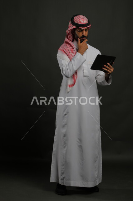 البحث عن معلومات مفيدة ومشاهدة البرامج التعليمية والتثقيفية, بورتريه لرجل عربي خليجي سعودي يرتدي الثوب التقليدي والشماغ يقوم باستخدام جهاز التابلت بإيماءات التفكير، استخدام التكنولوجيا والتقنيات الحديثة, صورة الجسم بالكامل, خلفية سوداء