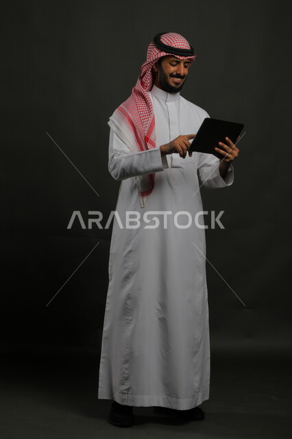 اللعب بالألعاب الالكترونية عبر الأجهزة الذكية, التواصل مع الاصدقاء عبر الاونلاين, بورتريه لرجل عربي خليجي سعودي مبتسم يرتدي الزي التقليدي يمسك جهاز التابلت بإيماءات السرور باستخدام التقنيات والتطبيقات الحديثة, صورة الجسم بالكامل, خلفية سوداء