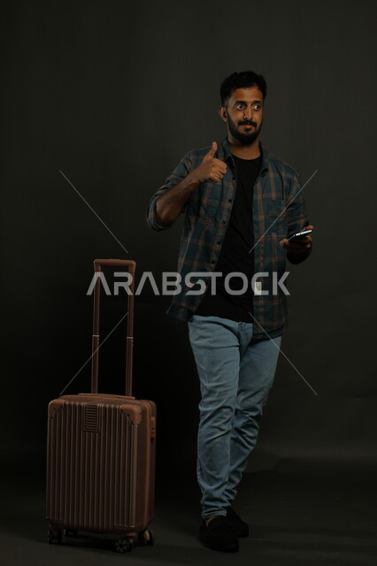 الاعجاب بخدمات العملاء ،الاستعداد لبدء الرحلة السياحية بإيماءات القبول والرضا، قضاء أوقات ممتعه خارج البلاد ، بورتريه لشاب عربي خليجي سعودي مبتسم يرتدي الزي الكاجوال يمسك بيده الجوال ويرفع اصبع الابهام للأعلى بعلامة الجودة، صورة للجسم بالكامل ، خلفية سوداء