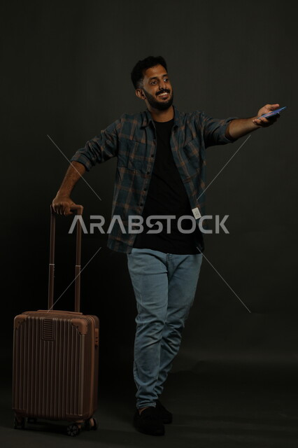 تأكيد الحجز الالكتروني عبر الجوال، بورتريه لشاب عربي خليجي سعودي يرتدي الزي الكاجوال يمسك بيده حقيبة السفر والهاتف المحمول، الاستعداد لبدء الرحلة في العطلة الصيفيه بحماس وسرور، صورة الجسم بالطول الكامل، خلفيه سوداء