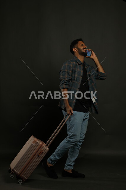 تبادل الأحاديث المسلية والمضحكة عبر الهاتف المحمول، إجراء مكالمة هاتفية عبر الجوال بتعابير الفرح، بورتريه لشاب عربي خليجي سعودي يرتدي الزي الكاجوال يقوم بجر حقيبة السفر خلفه ،ايماءات المشي والذهاب إلى المطار، صورة الجسم بالطول الكامل، خلفية سوداء