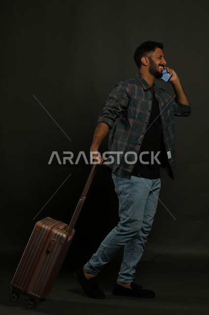 سماع الأخبار السارة قبل مغادرة البلاد، إجراء مكالمة هاتفية عبر الجوال بتعابير الفرح، بورتريه لشاب عربي خليجي سعودي يرتدي الزي الكاجوال يقوم بجر حقيبة السفر خلفه ،ايماءات المشي والذهاب إلى المطار، صورة الجسم بالطول الكامل، خلفية سوداء
