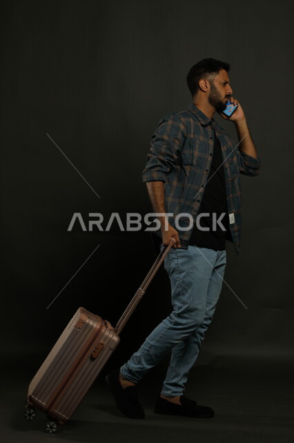 الاستفهام عن خدمات العملاء للرحلة السياحية, الاستفسار والسؤال عن أمر ما بإيماءات القلق,  بورتريه لرجل عربي خليجي سعودي يرتدي الزي الكاجوال يقوم بجر حقيبة السفر خلفه، إجراء مكالمة هاتفية عبر الهاتف المحمول, صورة الجسم بالطول الكامل, خلفية سوداء