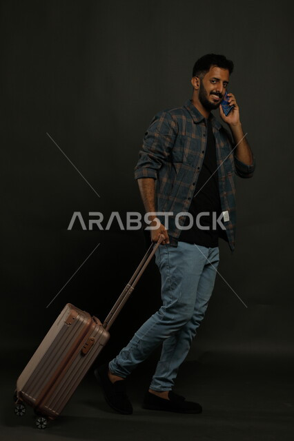 التحدث مع الأهل والأقارب قبل مغادرة البلاد، إجراء مكالمة هاتفية عبر الجوال والنظر إلى الكاميرا، بورتريه لشاب عربي خليجي سعودي يرتدي الزي الكاجوال يقوم بجر حقيبة السفر خلفه ،ايماءات المشي والذهاب إلى المطار، صورة الجسم بالطول الكامل، خلفية سوداء