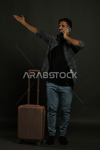 الاستقبال بشوق وسرور وحماس ,إجراء مكالمة هاتفية عبر الهاتف المحمول مع الاصدقاء، بورتريه لشاب عربي سعودي خليجي مبتسم يرتدي الزي الكاجوال يشير بيده بإيماءات السرور باللقاء في أحد الأقارب, صورة الجسم بالطول الكامل, خلفية رمادية