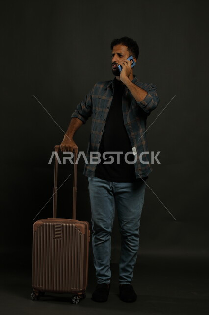 التواصل مع أحد موظفي الريسبشن للتأكد من الحجز, إجراء مكالمة هاتفية عبر الهاتف المحمول، بورتريه لشاب عربي سعودي خليجي يرتدي الزي الكاجوال يمسك بيده حقيبة السفر بإيماءات القلق, حجوزات مباشرة عبر تطبيقات وبرامج السياحة, صورة الجسم بالكامل, خلفية رمادية