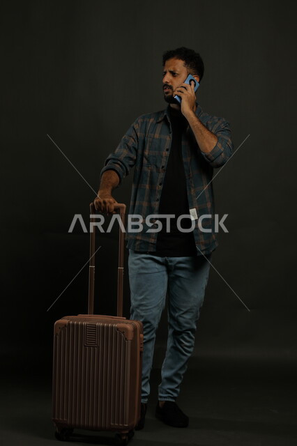 إجراء مكالمة هاتفية عبر الهاتف المحمول، التواصل مع أحد موظفي الريسبشن للتأكد من الحجز, بورتريه لشاب عربي سعودي خليجي مبتسم يرتدي الزي الكاجوال يمسك بيده حقيبة السفر بإيماءات القلق, حجوزات مباشرة عبر تطبيقات وبرامج السياحة, صورة الجسم بالكامل, خلفية رمادية