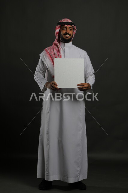 موك اب مربع أبيض، إعلانات وعروض تسويقية، استخدام لوحات توضيحية ووسائل تعليمية، بورتريه لشاب عربي خليجي سعودي مبتسم يرتدي الثوب التقليدي والشماغ يمسك بيديه لوحه بيضاء مربعه فارغة، صورة الجسم بالطول الكامل، خلفية سوداء