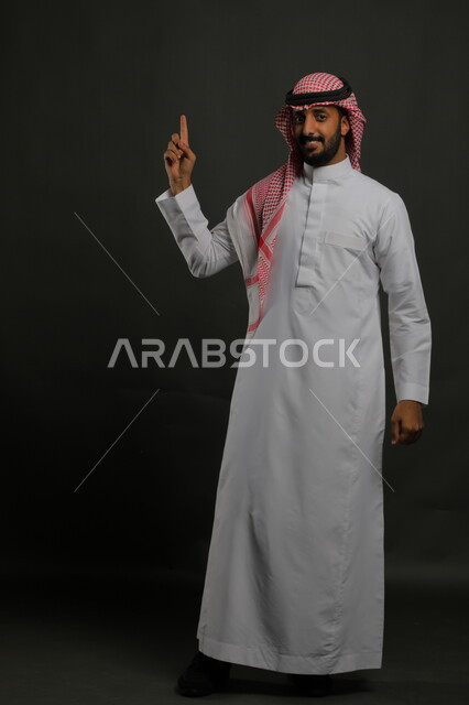 الإشارة الى الأعلى ، الوقوف باستقامة وانضباط،  الاهتمام بالمظهر الخارجي، بورتريه لرجل عربي خليجي سعودي يرتدي الزي التقليدي والشماغ وينظر إلى الكاميرا بابتسامة، مفهوم الاناقه والرجولة، صورة لكامل الجسم، خلفية رمادية