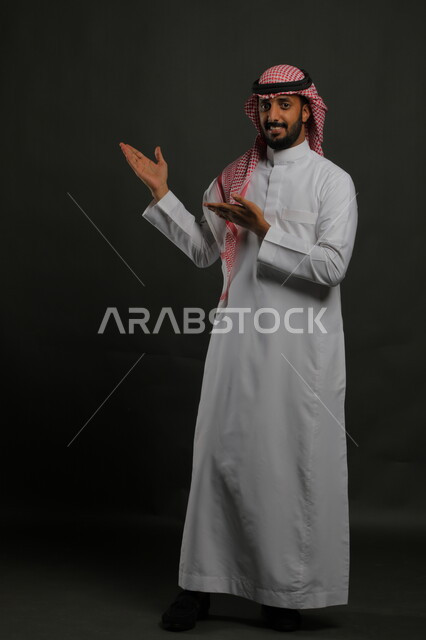 الإشارة الى مكان ما، الوقوف باستقامة وانضباط،  الاهتمام بالمظهر الخارجي، بورتريه لرجل عربي خليجي سعودي يرتدي الزي التقليدي والشماغ وينظر إلى الكاميرا بابتسامة، مفهوم الاناقه والرجولة، صورة لكامل الجسم، خلفية رمادية
