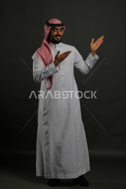 الإشارة الى مكان ما، الوقوف باستقامة وانضباط،  الاهتمام بالمظهر الخارجي، بورتريه لرجل عربي خليجي سعودي يرتدي الزي التقليدي والشماغ وينظر إلى الكاميرا بابتسامة، مفهوم الاناقه والرجولة، صورة لكامل الجسم، خلفية رمادية