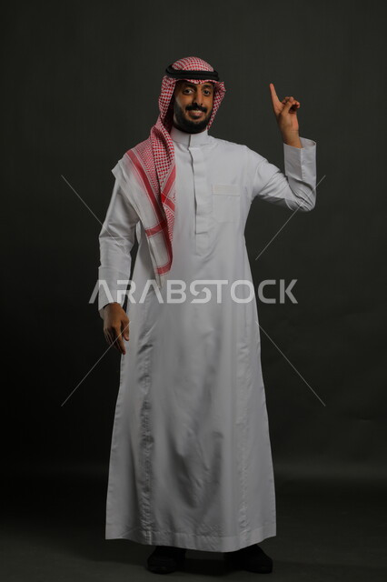 الإشارة الى الأعلى ، الوقوف باستقامة وانضباط،  الاهتمام بالمظهر الخارجي، بورتريه لرجل عربي خليجي سعودي يرتدي الزي التقليدي والشماغ وينظر إلى الكاميرا بابتسامة، مفهوم الاناقه والرجولة، صورة لكامل الجسم، خلفية رمادية