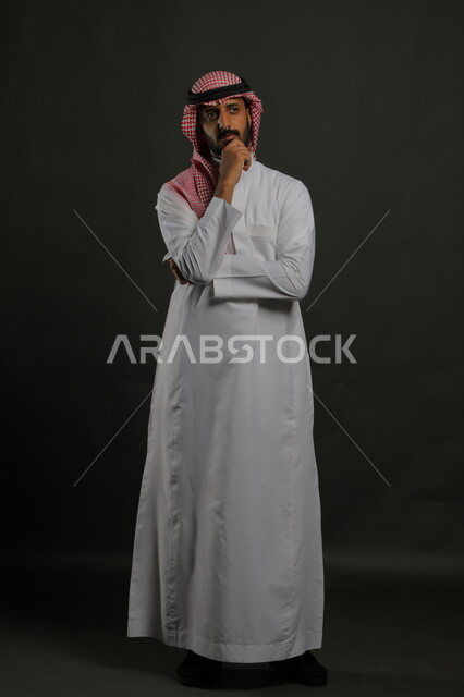 ايماءات التفكير والحيرة، الوقوف باستقامة وانضباط،  الاهتمام بالمظهر الخارجي، بورتريه لرجل عربي خليجي سعودي يرتدي الزي التقليدي والشماغ وينظر إلى الكاميرا بجدية، مفهوم الاناقه والرجولة، صورة لكامل الجسم، خلفية رمادية