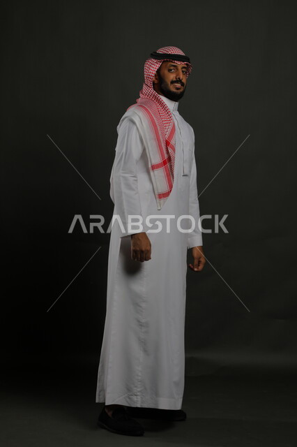 الثقة بالنفس والشعور بالقوة، الاهتمام بالمظهر الخارجي، بورتريه لرجل عربي خليجي سعودي يرتدي الزي التقليدي والشماغ وينظر إلى الكاميرا بجدية، مفهوم الاناقه والرجولة، الوقوف باستقامة وانضباط، صورة لكامل الجسم، خلفية رمادية
