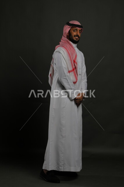 ايماءات الثقة والاعتزاز بالنفس، الاهتمام بالمظهر الخارجي، بورتريه لرجل عربي خليجي سعودي يرتدي الزي التقليدي والشماغ وينظر إلى الكاميرا بجدية، مفهوم الاناقه والرجولة، الوقوف باستقامة وانضباط، صورة لكامل الجسم، خلفية رمادية