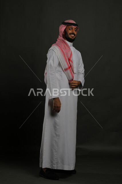ايماءات الثقة والاعتزاز بالنفس، الاهتمام بالمظهر الخارجي، بورتريه لرجل عربي خليجي سعودي يرتدي الزي التقليدي والشماغ وينظر إلى الكاميرا بابتسامة، مفهوم الاناقه والرجولة، الوقوف باستقامة وانضباط، صورة لكامل الجسم، خلفية رمادية
