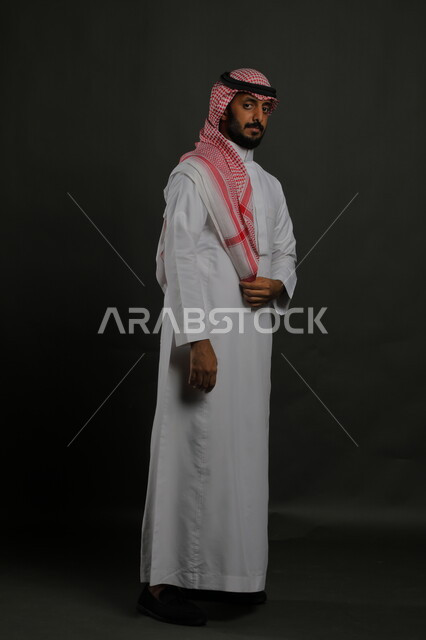 الوقوف باستقامة وثقه بالنفس، الاهتمام بالمظهر الخارجي، بورتريه لرجل عربي خليجي سعودي يرتدي الزي التقليدي والشماغ وينظر إلى الكاميرا بجدية، مفهوم الاناقه والرجولة، صورة لكامل الجسم، خلفية رمادية