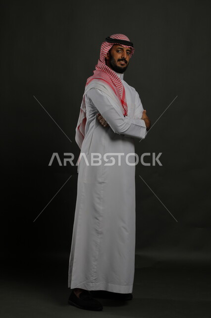  مفهوم الرجولة والأناقة، الاهتمام بالمظهر الخارجي، بورتريه لشاب عربي خليجي سعودي يرتدي الزي التقليدي والشماغ يقف مكتف اليدين وينظر الى الكاميرا بايماءات الثقة والاعتزاز بالنفس،  صورة لكامل الجسم، خلفية رمادية
