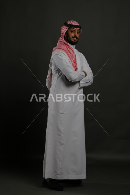  مفهوم الاناقه والرجولة، الاهتمام بالمظهر الخارجي، بورتريه لرجل عربي خليجي سعودي يرتدي الزي التقليدي والشماغ يقف بأيدي متقاطعة وينظر الى الكاميرا بايماءات الثقة والاعتزاز بالنفس، صورة الجسم بالطول الكامل، خلفية رمادية