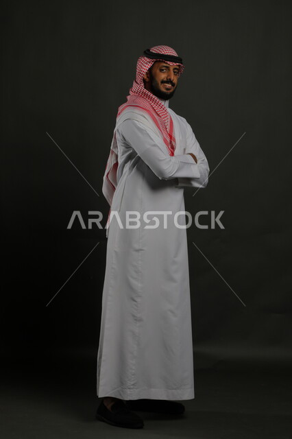  مفهوم الاناقه والرجولة، الاهتمام بالمظهر الخارجي، بورتريه لرجل عربي خليجي سعودي يرتدي الزي التقليدي والشماغ يقف مكتف اليدين وينظر الى الكاميرا بايماءات الثقة والاعتزاز بالنفس،  صورة لكامل الجسم، خلفية رمادية
