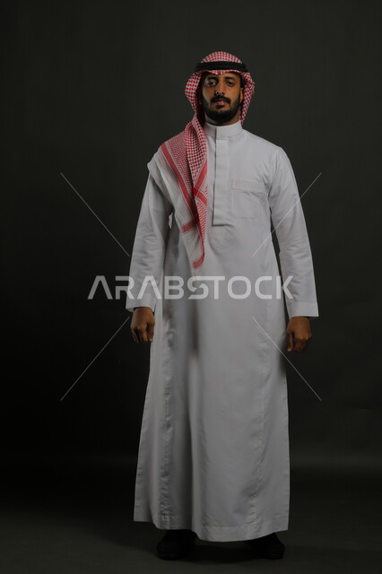  الاهتمام بالمظهر الخارجي، بورتريه لشاب عربي خليجي سعودي يرتدي الزي التقليدي والشماغ وينظر إلى الكاميرا بإيماءات الجدية و الثقة بالنفس، مفهوم الاناقه والرجولة، صورة الجسم بالطول الكامل، خلفية رمادية