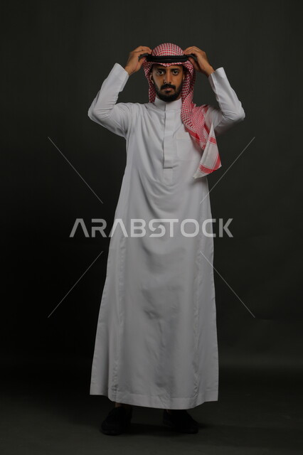  الاهتمام بالمظهر الخارجي، بورتريه لرجل عربي خليجي سعودي يرتدي الزي التقليدي والشماغ وينظر إلى الكاميرا بإيماءات الجدية و الثقة بالنفس، مفهوم الاناقه والرجولة، صورة لكامل الجسم، خلفية رمادية