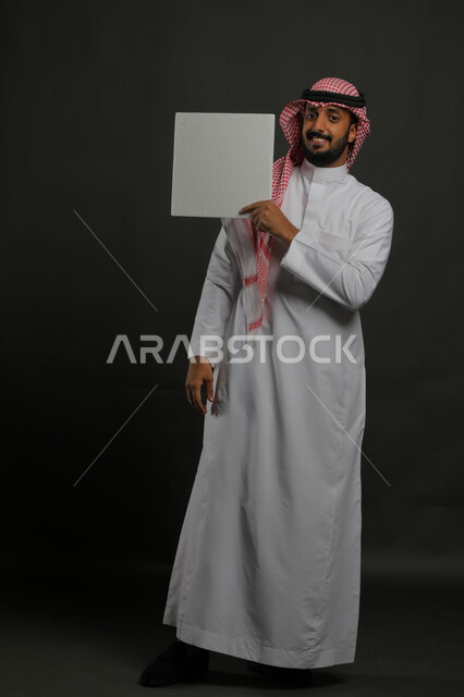 موك اب مربع أبيض، إعلانات وعروض تسويقية، استخدام لوحات توضيحية ووسائل تعليمية، بورتريه لشاب عربي خليجي سعودي مبتسم يرتدي الثوب التقليدي والشماغ يمسك بيده لوحه بيضاء مربعه فارغة، صورة الجسم بالطول الكامل، خلفية سوداء