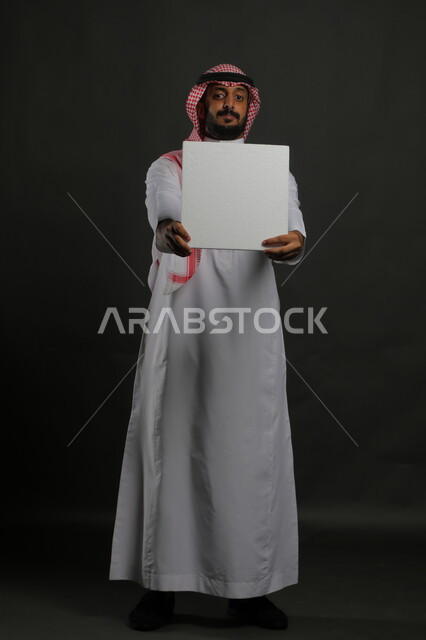 عروض وإعلانات تسويقية ترويجية، موك اب مربع أبيض، استخدام لوحات توضيحية ووسائل تعليمية، بورتريه لشاب عربي خليجي سعودي يرتدي الثوب التقليدي والشماغ يمسك بيديه لوحه بيضاء مربعه فارغة، صورة الجسم بالطول الكامل، خلفية سوداء