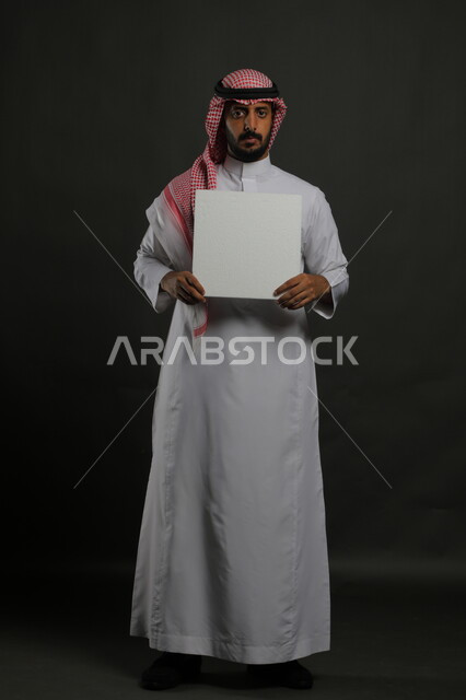 النظر إلى الكاميرا بإيماءات الثقة، عروض وإعلانات تسويقية ترويجية، استخدام لوحات توضيحية ووسائل تعليمية، بورتريه لرجل عربي خليجي سعودي يرتدي الثوب التقليدي والشماغ يمسك بيديه موك اب مربع أبيض، لوحه بيضاء مربعه فارغة، صورة الجسم بالطول الكامل، خلفية سوداء