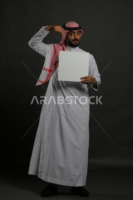 الإشارة إلى لوحة بيضاء مربعه فارغة، العروض والإعلانات التسويقية الترويجية، استخدام اللوحات التوضيحية والوسائل التعليمية التقليدية، بورتريه لرجل عربي خليجي سعودي مبتسم يرتدي الثوب التقليدي والشماغ يمسك بيديه موك اب مربع أبيض، صورة الجسم بالكامل، خلفية سوداء