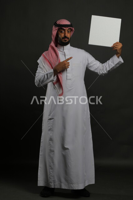 الإشارة إلى لوحة بيضاء مربعه فارغة، العروض والإعلانات التسويقية الترويجية، استخدام اللوحات التوضيحية والوسائل التعليمية التقليدية، بورتريه لرجل عربي خليجي سعودي مبتسم يرتدي الثوب التقليدي والشماغ يمسك بيديه موك اب مربع أبيض، صورة الجسم بالكامل، خلفية سوداء