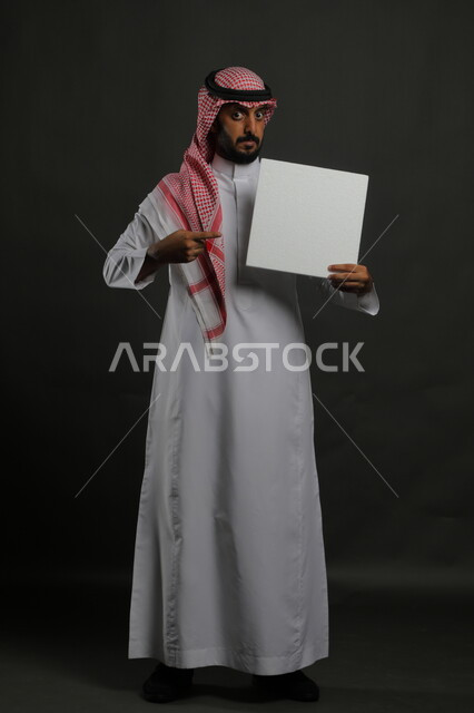 الإشارة إلى لوحة بيضاء مربعه فارغة، العروض والإعلانات التسويقية الترويجية، استخدام اللوحات التوضيحية والوسائل التعليمية التقليدية، بورتريه لرجل عربي خليجي سعودي مبتسم يرتدي الثوب التقليدي والشماغ يمسك بيديه موك اب مربع أبيض، صورة الجسم بالكامل، خلفية سوداء