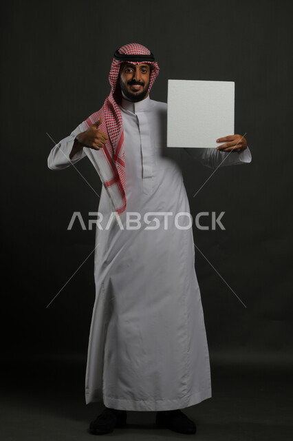 الإشارة بعلامة الجودة والإعجاب، النظر إلى الكاميرا بإيماءات القبول والرضا، لوحة بيضاء مربعه فارغة، العروض والإعلانات التسويقية ، بورتريه لرجل عربي خليجي سعودي مبتسم يرتدي الثوب التقليدي والشماغ يمسك بيديه موك اب مربع أبيض، صورة الجسم بالكامل، خلفية سوداء