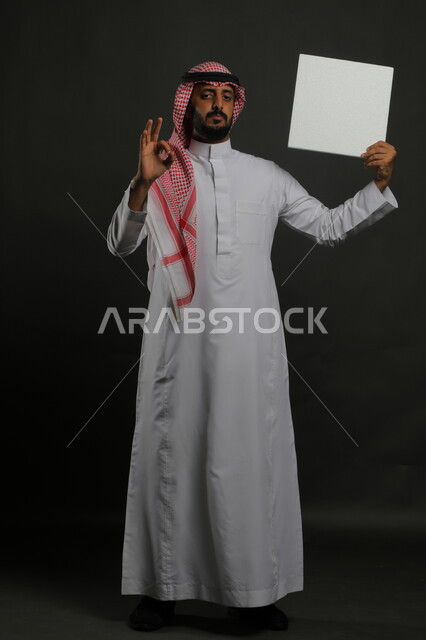 العروض والإعلانات التسويقية، الإشارة بعلامة الجودة والإعجاب، النظر إلى الكاميرا بإيماءات القبول والموافقة، لوحة بيضاء مربعه فارغة، بورتريه لرجل عربي خليجي سعودي يرتدي الثوب التقليدي والشماغ يمسك بيديه موك اب مربع أبيض، صورة الجسم بالكامل، خلفية سوداء
