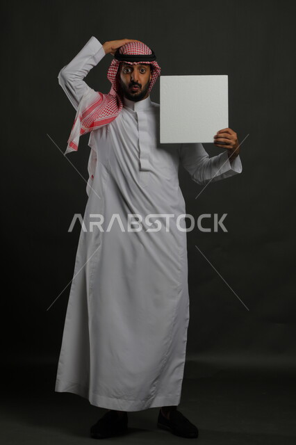 عرض لوحة بيضاء مربعه فارغة، العروض والإعلانات التسويقية، النظر إلى الكاميرا بإيماءات الدهشة والصدمة، بورتريه لرجل عربي خليجي سعودي يرتدي الثوب التقليدي والشماغ يمسك بيديه موك اب مربع أبيض، صورة الجسم بالكامل، خلفية سوداء