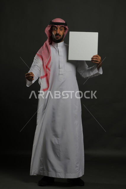 عرض لوحة بيضاء مربعه فارغة، العروض والإعلانات التسويقية، النظر إلى الكاميرا بإيماءات الدهشة والصدمة، بورتريه لرجل عربي خليجي سعودي يرتدي الثوب التقليدي والشماغ يمسك بيديه موك اب مربع أبيض، صورة الجسم بالكامل، خلفية سوداء