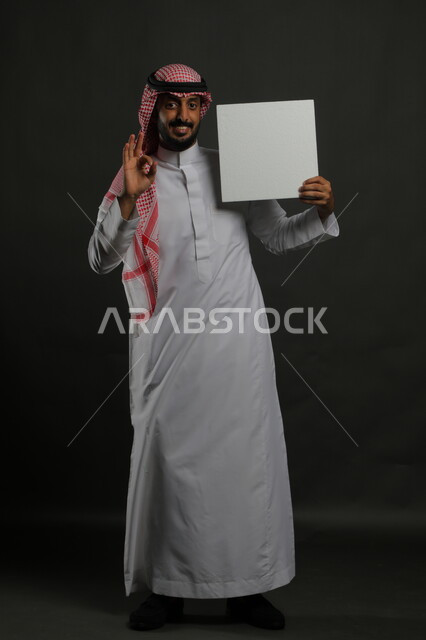 الإشارة بعلامة الجودة والامتياز، النظر إلى الكاميرا بإيماءات القبول والرضا، عرض لوحة بيضاء مربعه فارغة، العروض والإعلانات التسويقية ، بورتريه لرجل عربي خليجي سعودي مبتسم يرتدي الثوب التقليدي والشماغ يمسك بيديه موك اب مربع أبيض، صورة الجسم بالكامل، خلفية سوداء