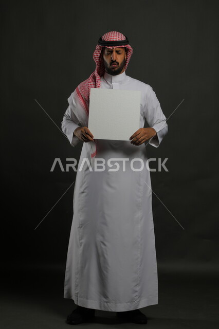لوحات توضيحية ووسائل تعليمية تقليدية، إيماءات الغضب والانزعاج، عرض لوحة بيضاء مربعه فارغة، بورتريه لرجل عربي خليجي سعودي يرتدي الثوب التقليدي والشماغ يمسك بيديه موك اب مربع أبيض، صورة الجسم بالكامل، خلفية سوداء