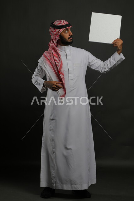 لوحات توضيحية ووسائل تعليمية تقليدية، عرض لوحة بيضاء مربعه فارغة، بورتريه لرجل عربي خليجي سعودي يرتدي الثوب التقليدي والشماغ يمسك بيديه موك اب مربع أبيض، صورة الجسم بالكامل، خلفية سوداء