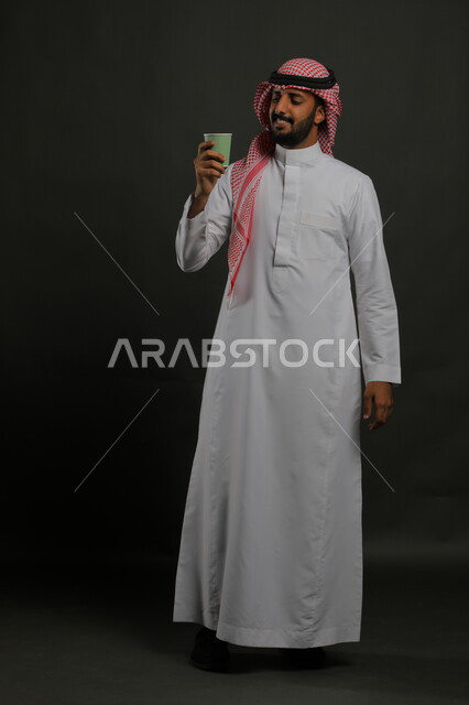 الاستمتاع بمذاق القهوة العربية، بورتريه لرجل عربي خليجي سعودي مبتسم يرتدي الزي السعودي التقليدي يحمل بيده كوب من الورق المقوى, إحتساء المشروبات الساخنه, صورة الجسم بالطول الكامل، خلفية سوداء