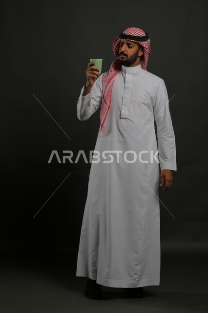 احتساء المشروبات الساخنه, الاستمتاع بمذاق القهوة العربية، بورتريه لرجل عربي خليجي سعودي يرتدي الزي السعودي التقليدي يحمل بيده كوب من الورق المقوى, صورة الجسم بالطول الكامل، خلفية سوداء