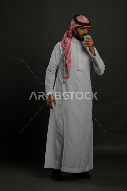 تعديل المزاج بتناول الكافيين، احتساء المشروبات الساخنه والأعشاب الشتوية، بورتريه لرجل عربي خليجي سعودي يرتدي الزي السعودي التقليدي يقوم بشرب القهوة العربية, صورة الجسم بالطول الكامل، خلفية سوداء