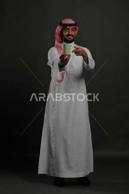 احتساء المشروبات الساخنه والأعشاب الشتوية الدافئة، الاستمتاع بتناول القهوة العربية، تعديل المزاج بتناول الكافيين، بورتريه لرجل عربي خليجي سعودي مبتسم يرتدي الزي السعودي التقليدي يشير بيده إلى كوب من الورق المقوى, صورة الجسم بالطول الكامل، خلفية سوداء
