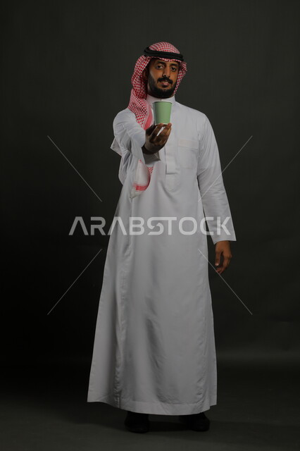 تأثير الكافيين على نشاط الدماغ والذاكرة، الاستمتاع بتناول المشروبات الساخنة والأعشاب الشتوية الدافئة، بورتريه لرجل عربي خليجي سعودي يرتدي الثوب التقليدي والشماغ يحمل بيده كوب ورقي مقوى من القهوة، صورة الجسم بالكامل، خليفية سوداء