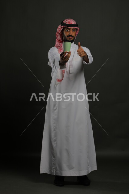 النظر إلى الكاميرا بإيماءات القبول والاستمتاع بتناول المشروبات الساخنة، بورتريه لرجل عربي خليجي سعودي يرتدي الثوب التقليدي والشماغ يحمل بيده كوب من القهوة ويشير بإصبع الإبهام بعلامة الجودة والإعجاب، صورة الجسم بالكامل، خليفية سوداء